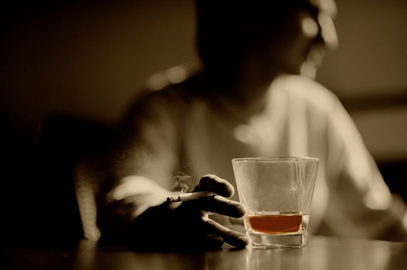 A man smoking and reaching for a glass of whiskey.