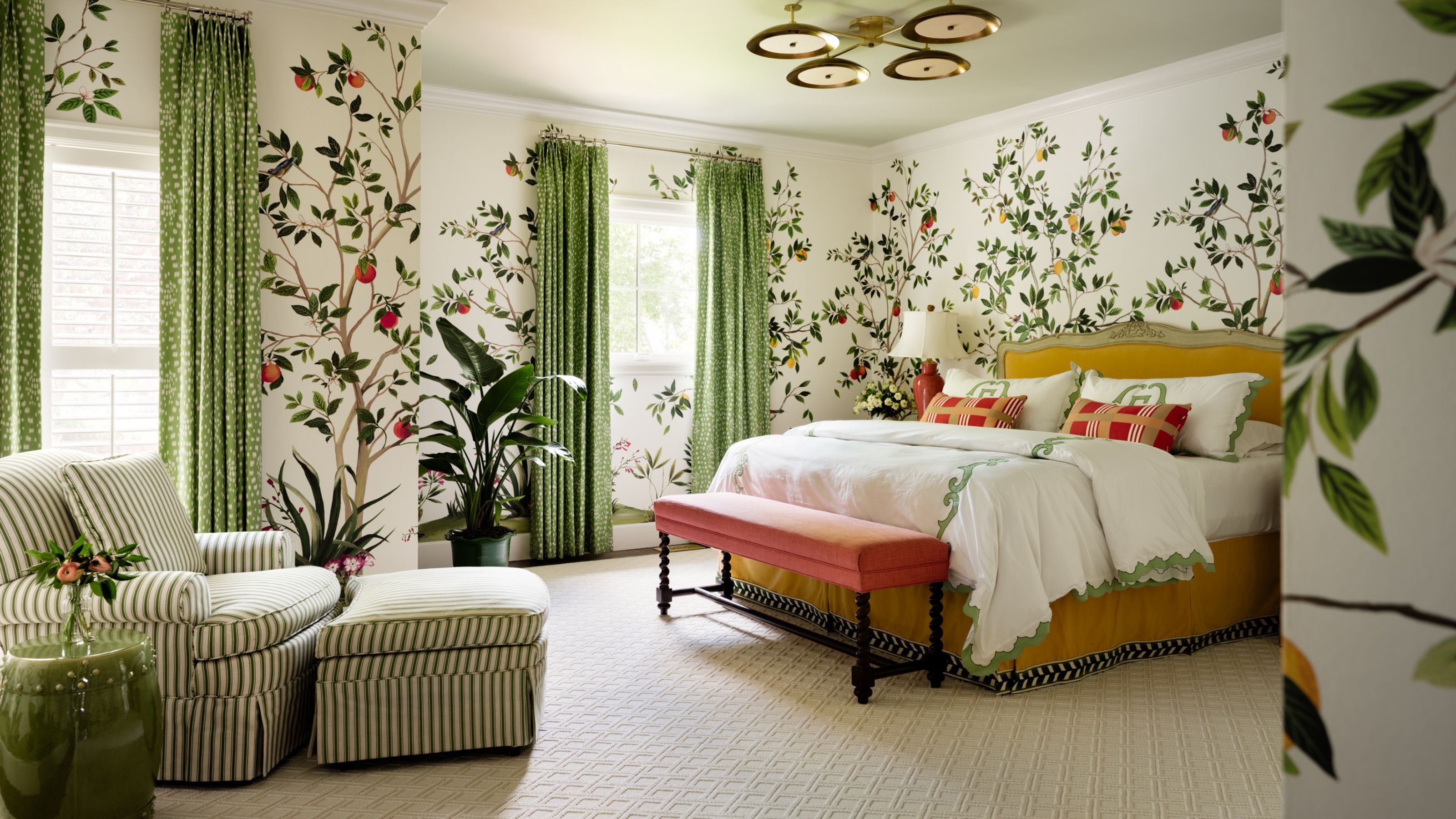 a neutral bedroom wrapped in botanical mural wallpaper with green drapes, a striped chair and stool and a yellow upholstered bed
