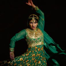 Priya Kansara does a martial arts pose in a green and gold dress against a black background