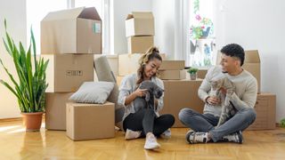Couple sat on floor in new house with their two cats surrounded by moving boxes