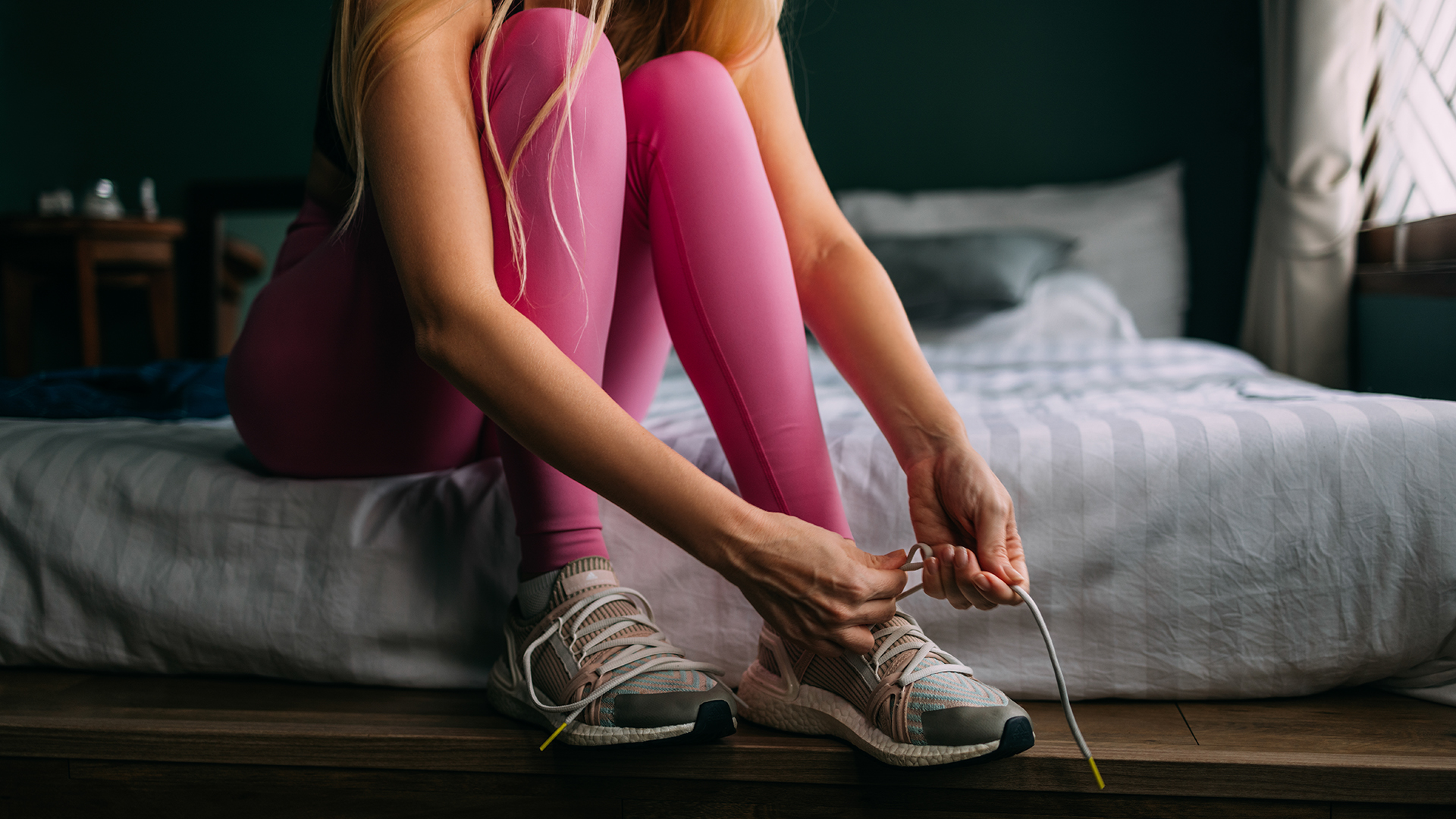 A woman sits on the corner of her bed wearing workout clothes and tying the laces on her running shoes