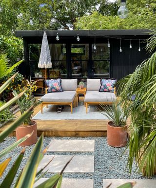 Black summerhouse with decking in front, two loungers, rug, umbrella, paved path, tropical plants