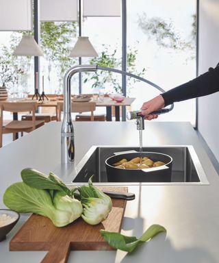 A modern kitchen with a sleek chrome pull-out tap filling a pan with potatoes. A wooden board with fresh pak choi sits beside a deep stainless steel sink.