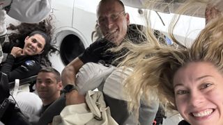 four people in blue flight suits laugh and smile as they float in zero gravity