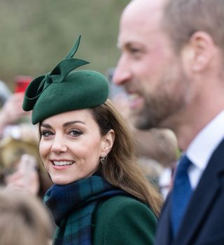 Kate Middleton wearing a green coat and hat, plaid scarf and smiling at Prince William who is blurred in the foreground