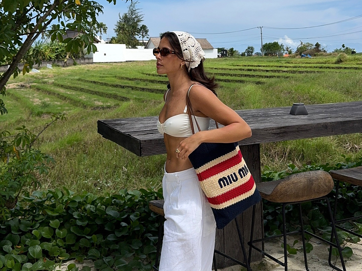 Influencer Tia Dewitt wearing a Miumiu knit tote, linen white pants, a white bikini top, and rope sandals.