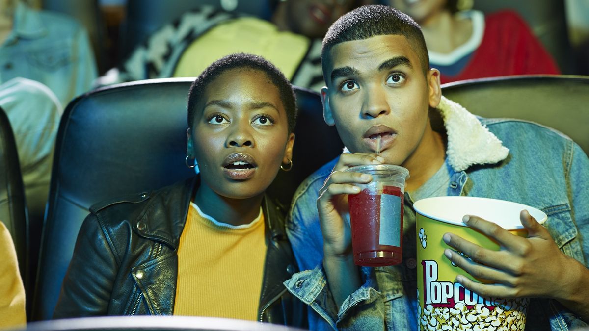 Two young adults lean together as they&#039;re watching a movie in a theater; one is holding a popcorn bucket and softdrink