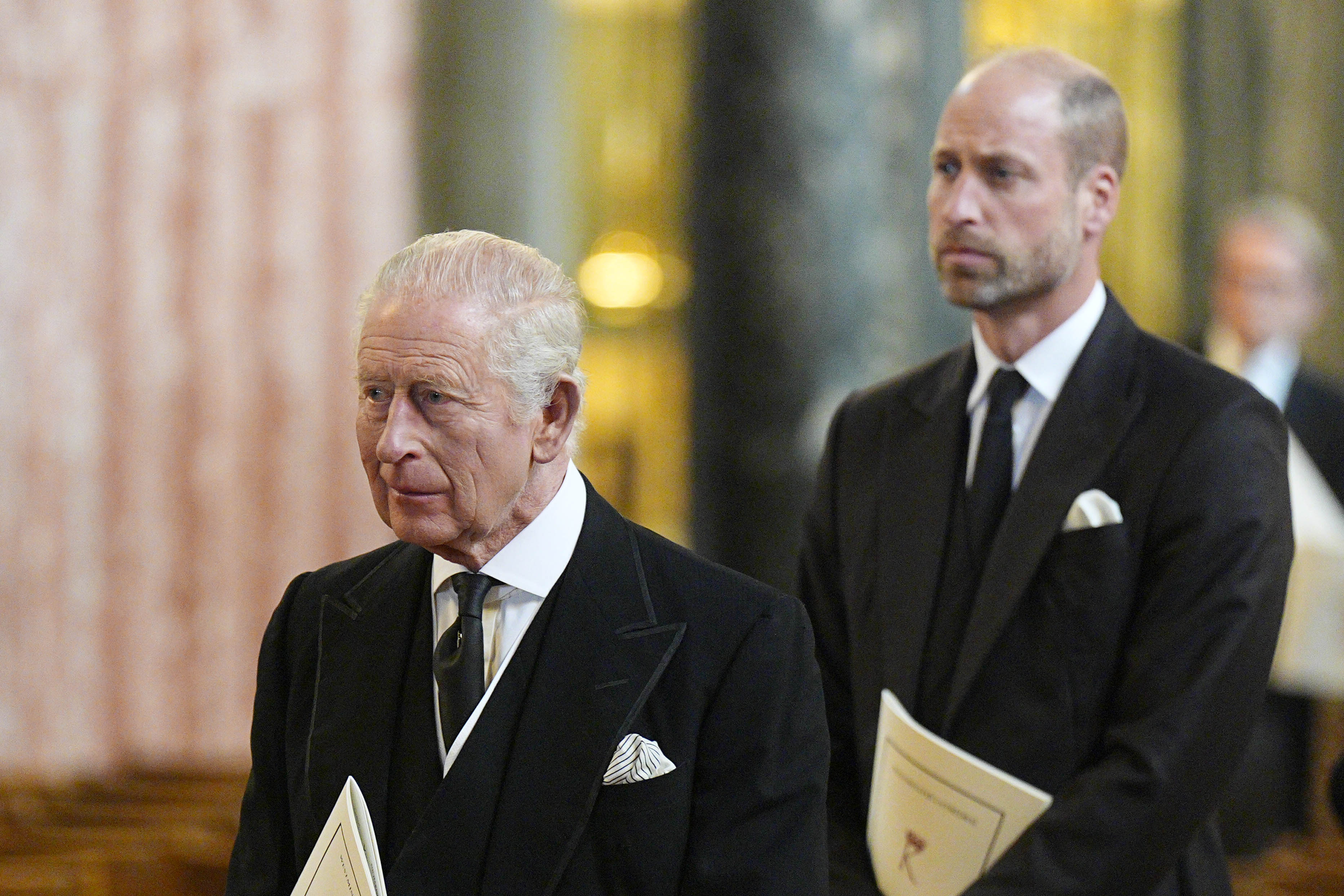 King Charles and Prince William wearing black suits holding programs