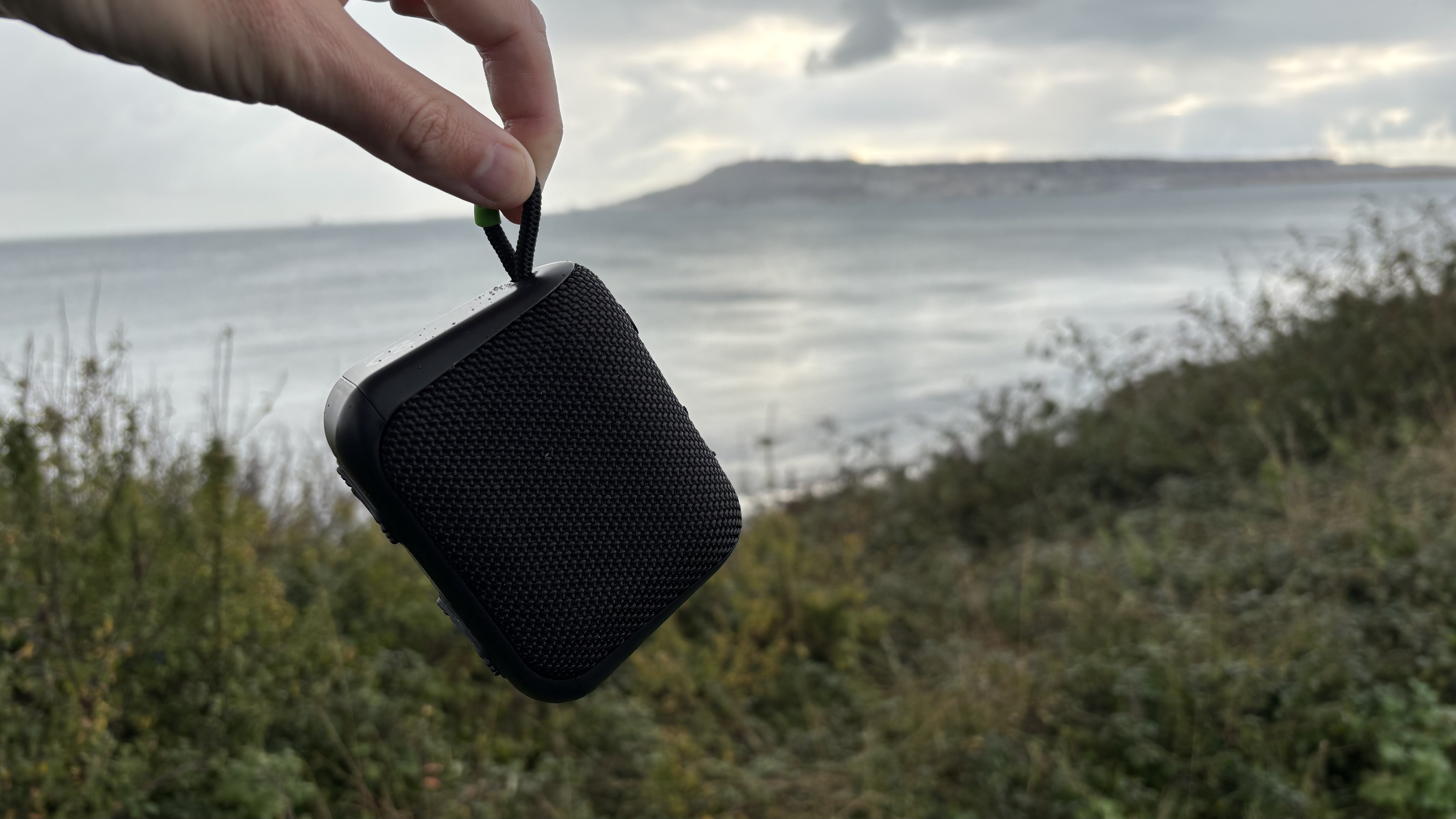 A hand holding the black AO mini portable wireless speaker on a raised promontory, with an island in the background.