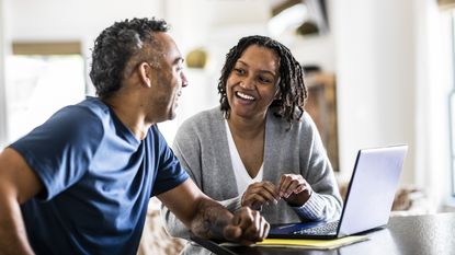 An older couple look at a laptop together at their kitchen table.