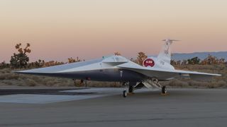 a white jet with an elongated nose section sits on a tarmac at sunset