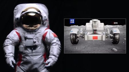 a white space suit with a gold visor next to a four-wheeled, car-like rover on a grey, dusty surface