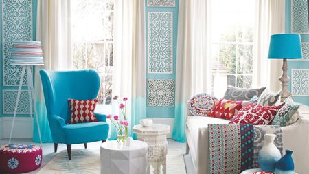 Bright and colorful patterned living room with blue walls, a blue chair, white couch, blue lamp, white coffee table with flowers, and patterned blankets, cushions, pouffe, table, lamp shade and vases.