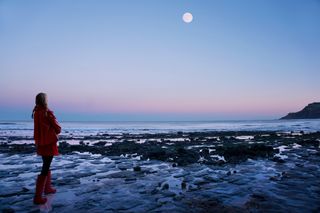 A woman weating a red coat stands on a beach with her back to the camera. A full moon shines high in the sky above the sea.