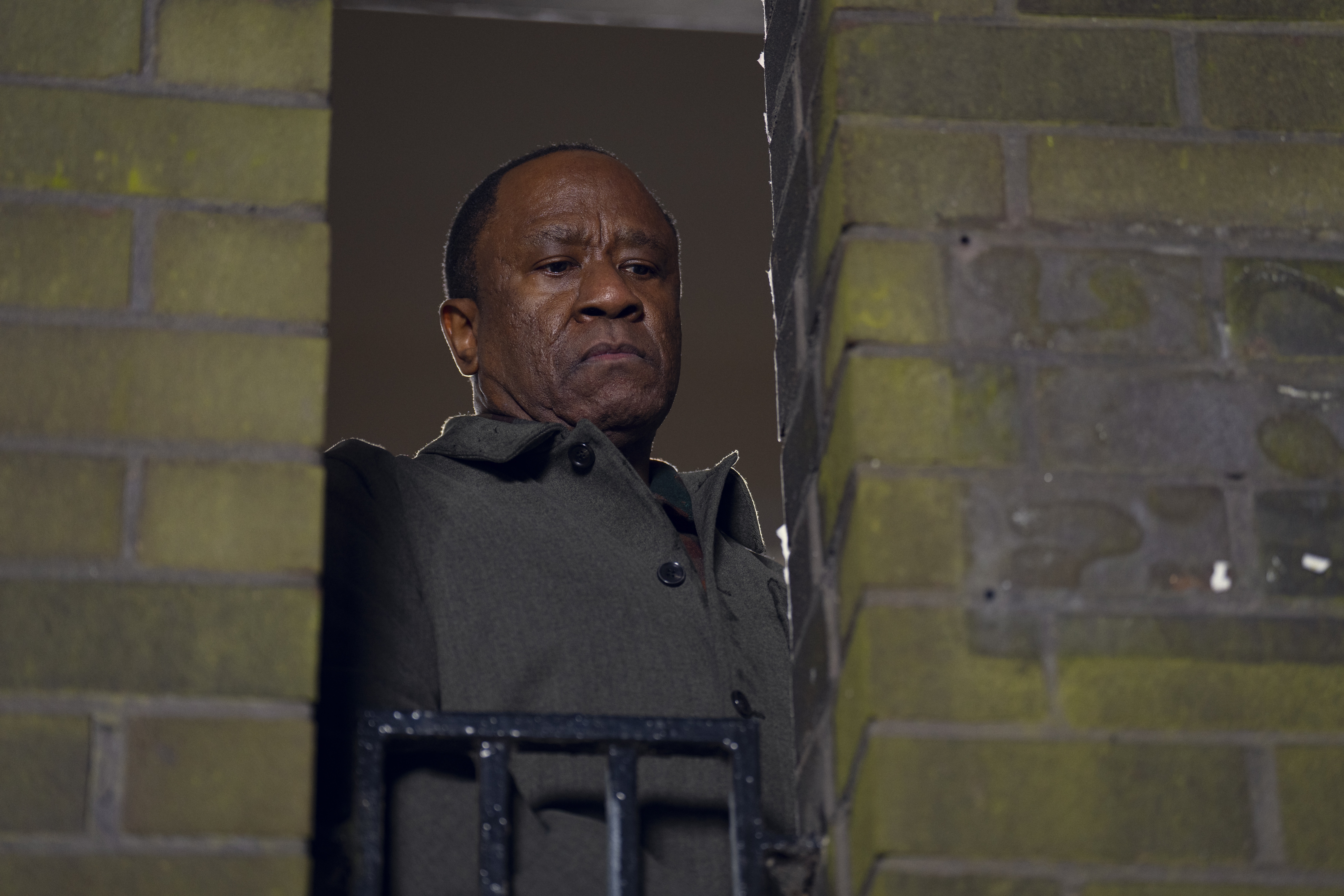 Lucien Msamati as cornelius faber wearing a grey peacoat looking out a brick window in a still from netflix's Run Away