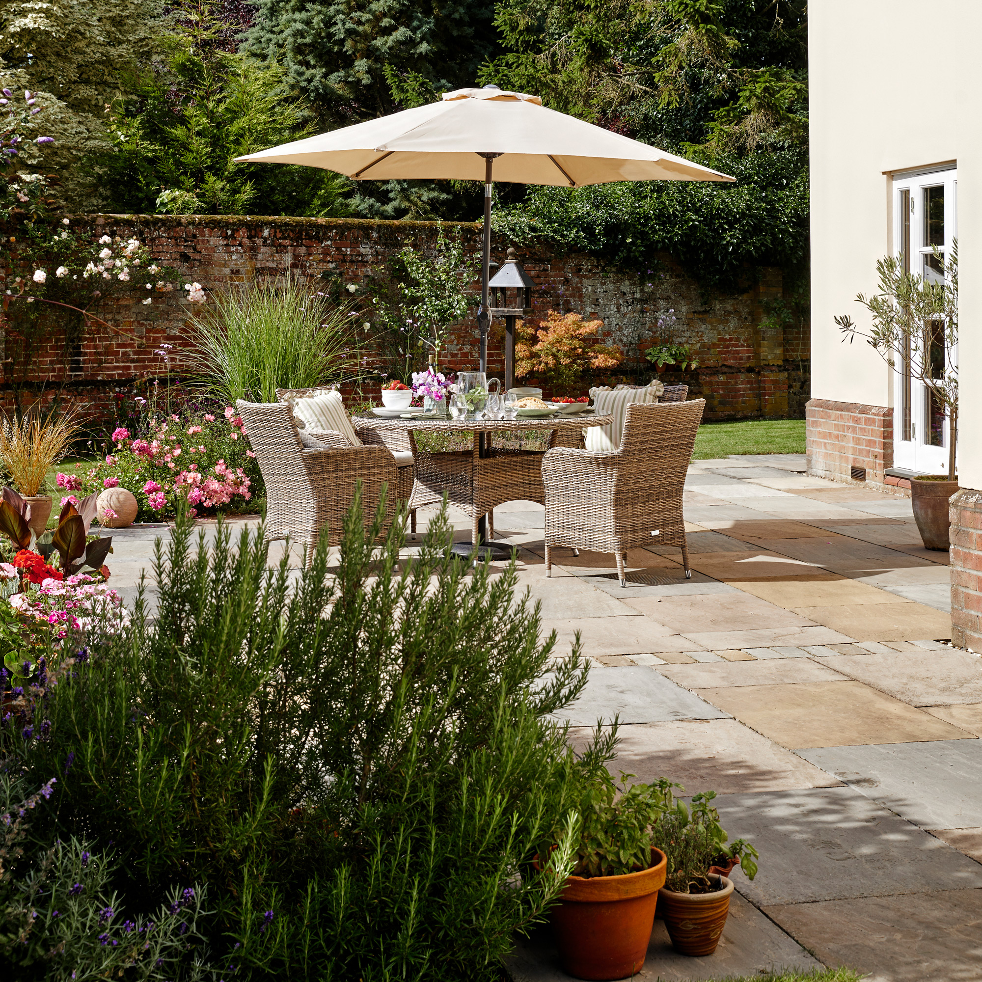 a patio with dining furniture and parasol alongside a garden wall with a large rosemary plant in the foreground