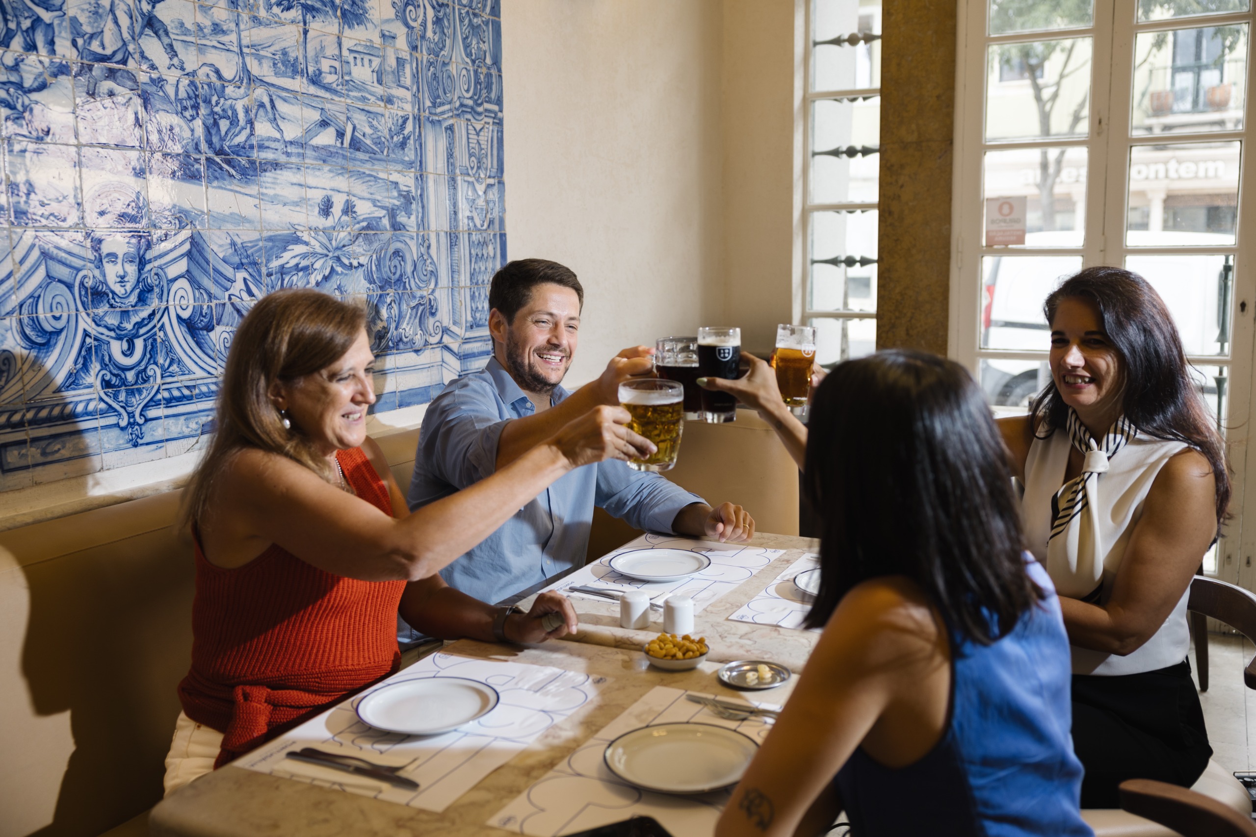 Four friends clink glasses of lager and dark beer across a marble table beside a large panel of blue-and-white painted tiles, with sunlight streaming through tall windows behind them.
