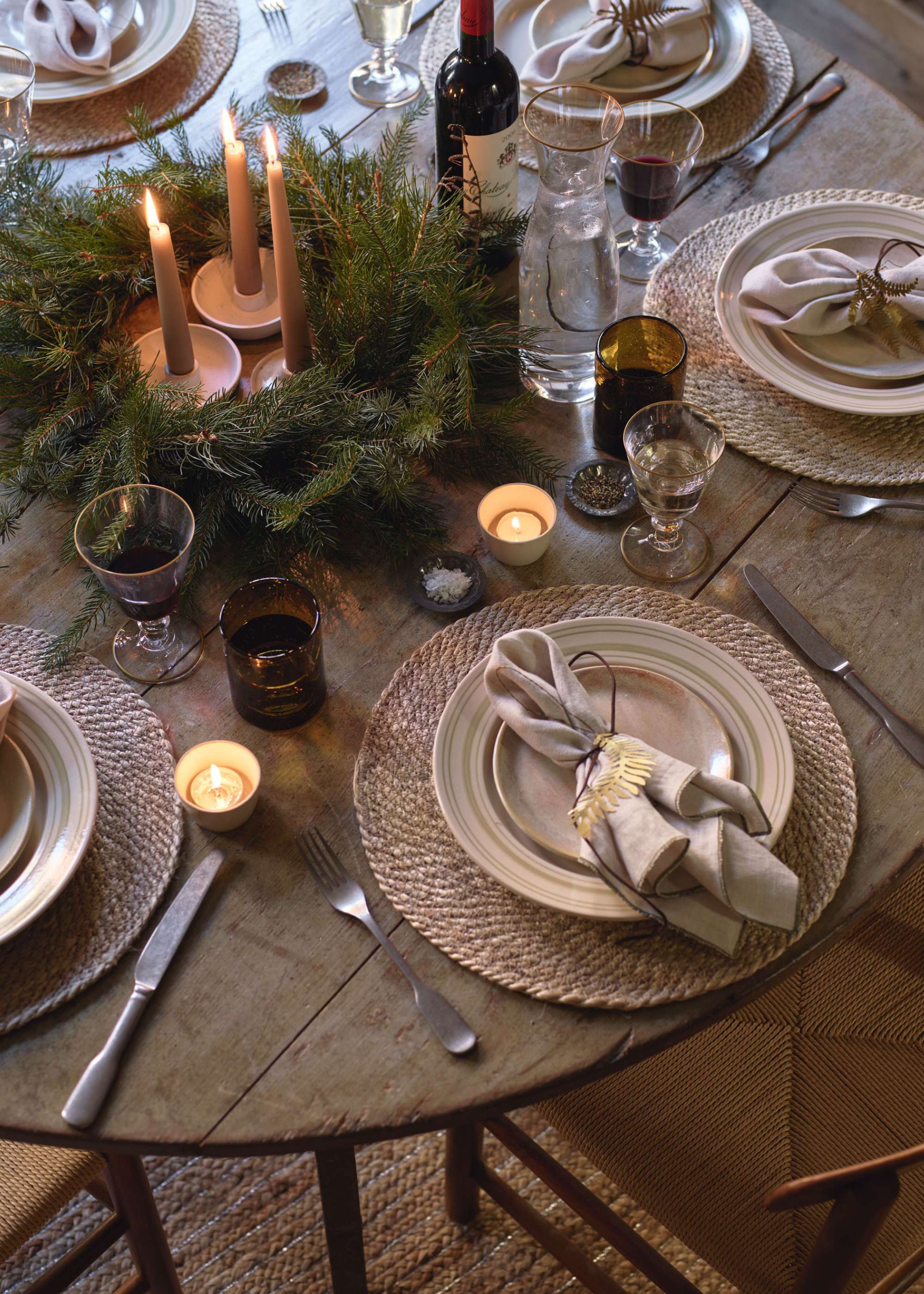 A round, wooden dining table set and decorated for Christmas with taper candles, tealights and foliage