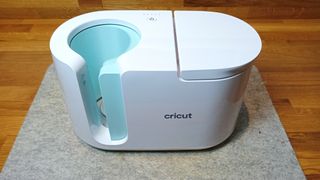 Best Cricut machines; an elogated white craft machine with rounded ends, there's a hole to place a round mug, it's on a grey heat mat on a wooden table