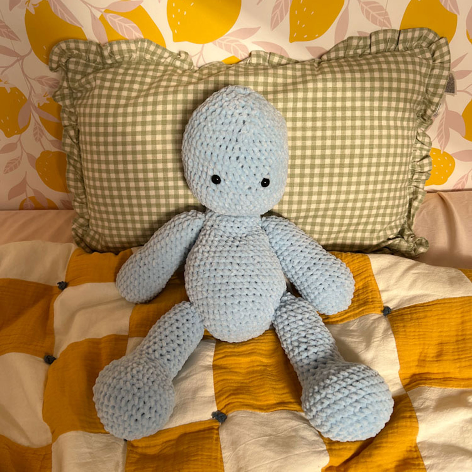 Blue knitted soft toy resting on green and white gingham cushion on top of yellow and white checked bedding
