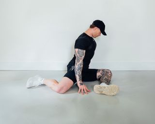 Coach Corey Perkins performs a 90/90 hip stretch with an upright torso. His left leg is in front of him, bent at a 90-degree angle, so that his calf is parallel. His right leg is also at a 90-degree angle, with his right knee held out to the side. His hands rest on the floor.