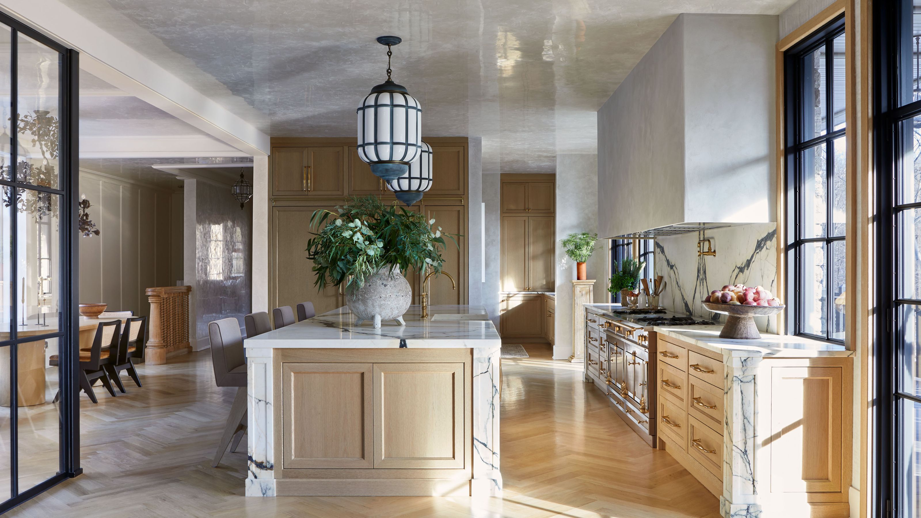 A large, open-plan kitchen with modern decor. Parquet flooring, a glossy ceiling, and a marble wall, kitchen island, and countertops. Pale wood cabinets and two black and glass pendant lights.