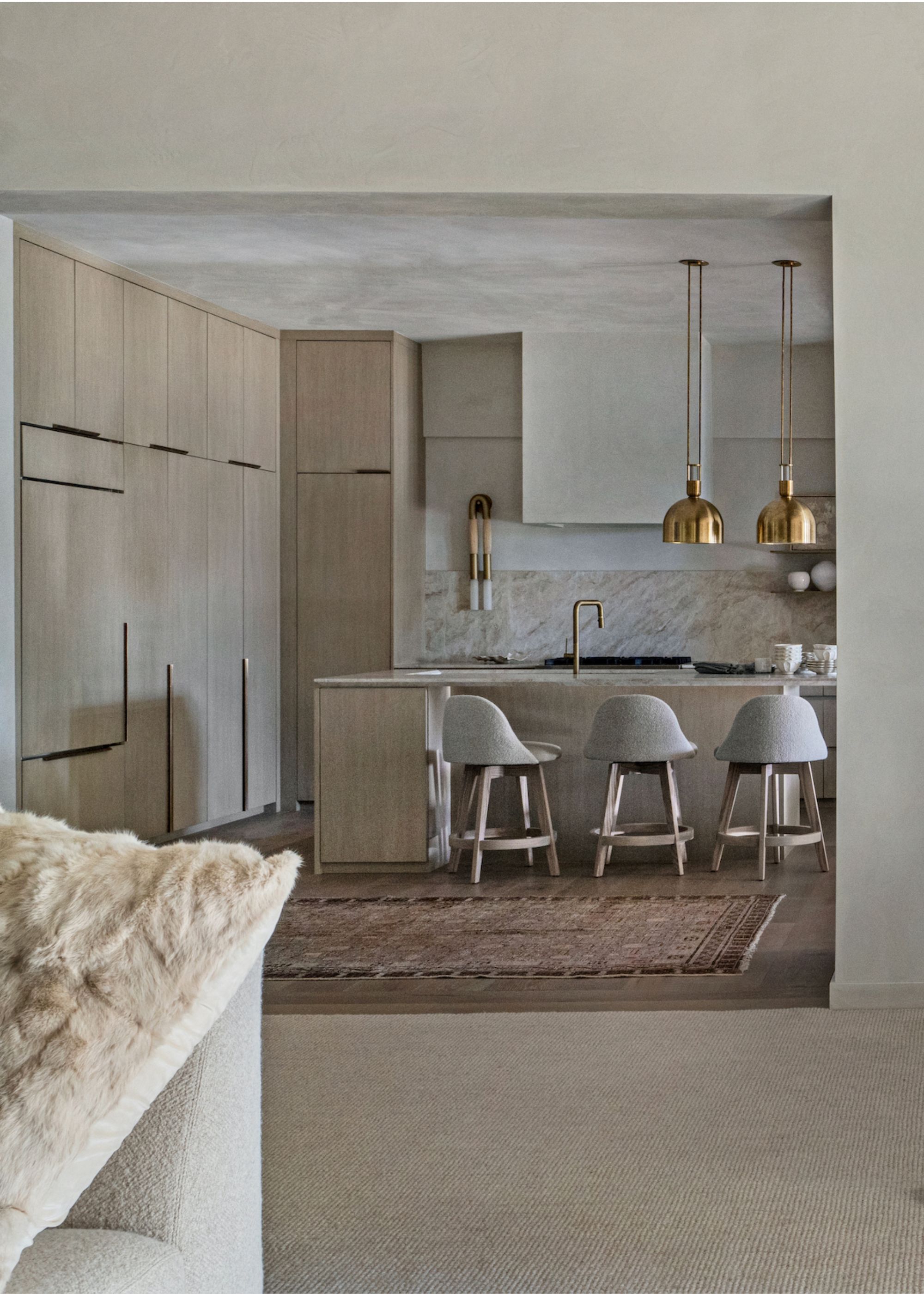 A living room cum kitchen, with the flooring fully carpeted, and kitchen designed with brass lights, and three bar chairs