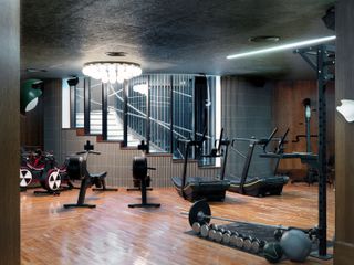 Interior view of the gym at White City House featuring wood flooring and walls, a dark coloured roof with strip and multi-sphere lighting, exercise equipment and glass windows offering a view of the stairwell outside the gym