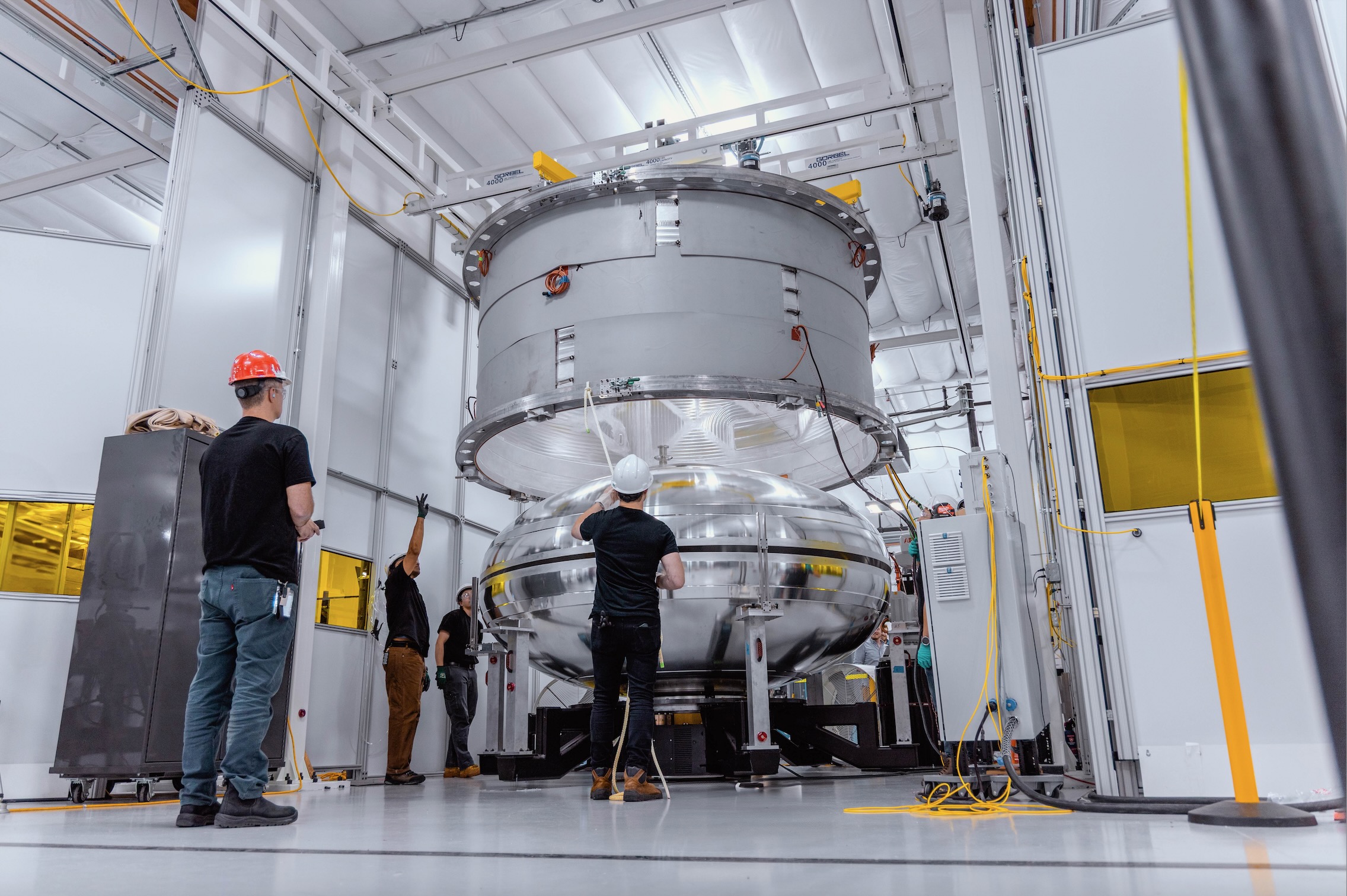 four people in hard hats stand around a shiny silver cylinder inside a white-walled room