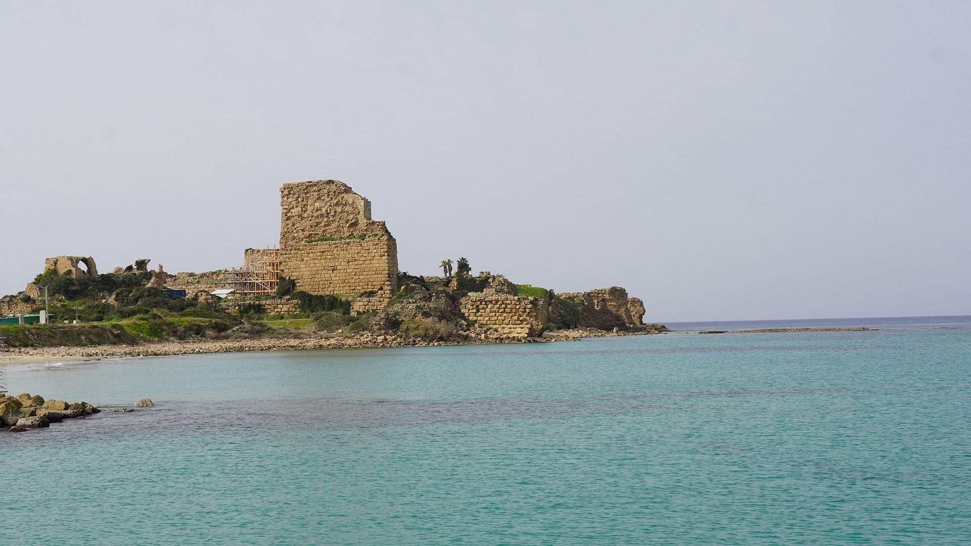A crumbling wall stands next to a beach with dark blue waters next to it.