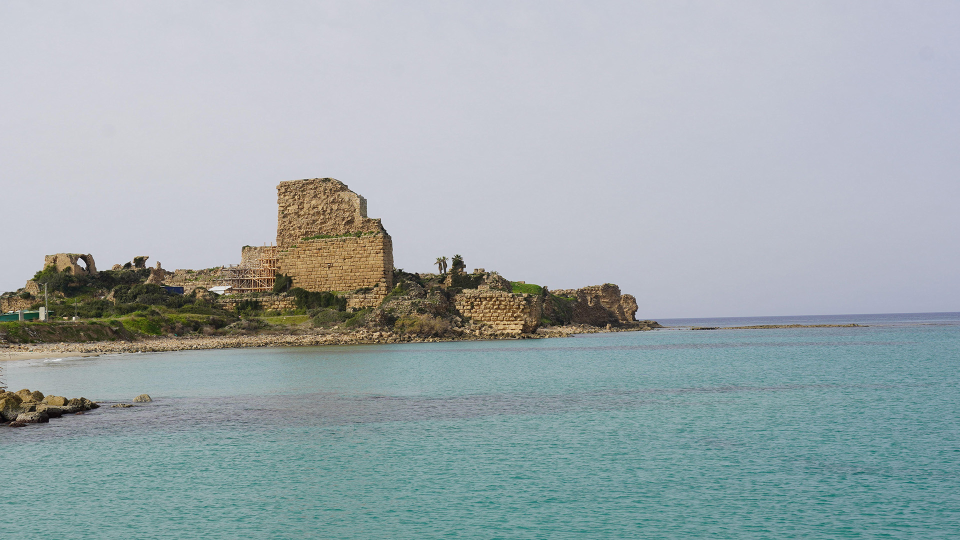 A crumbling wall stands next to a beach with dark blue waters next to it.