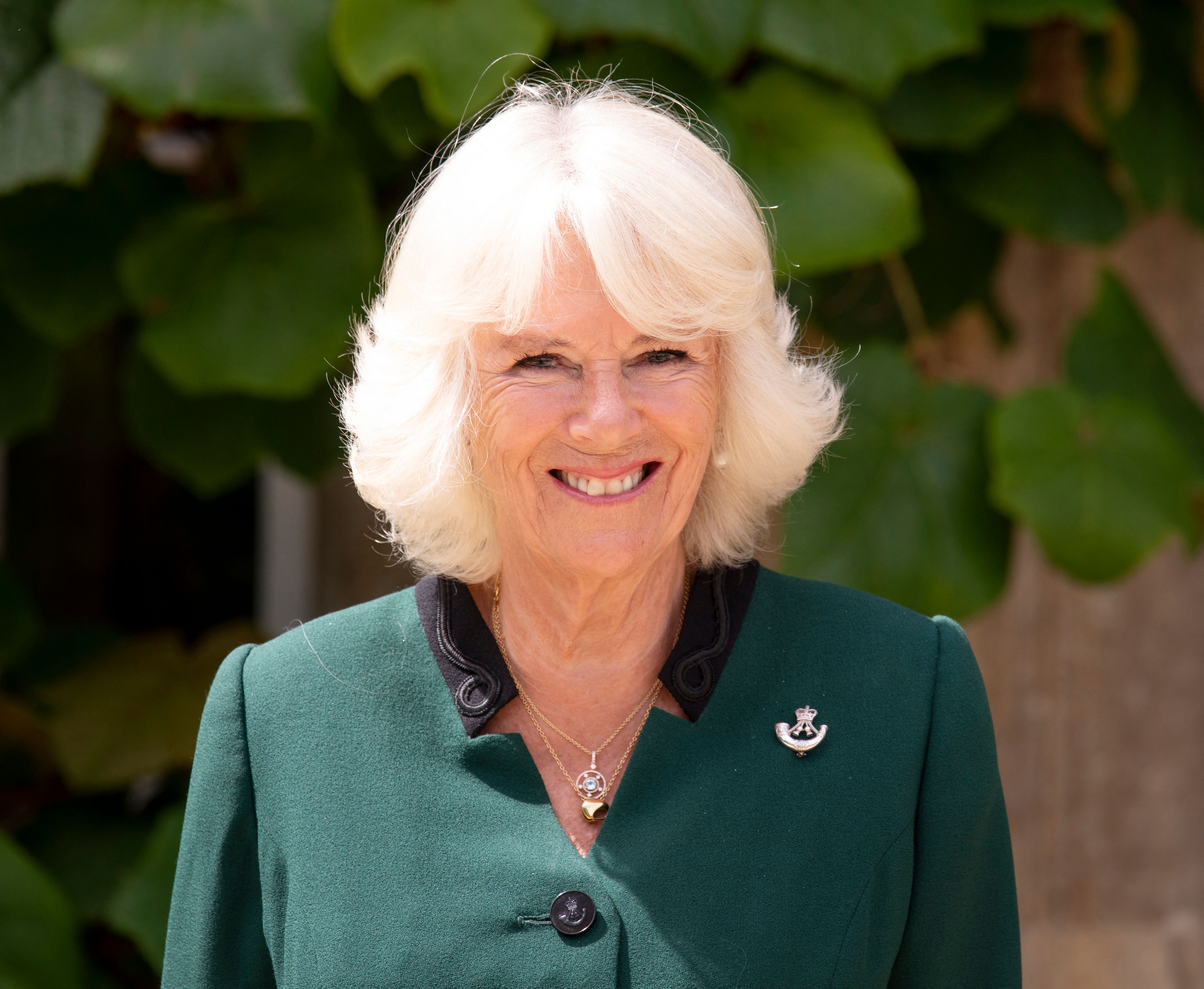 Queen Camilla wearing a green coat and smiling in front of a tree