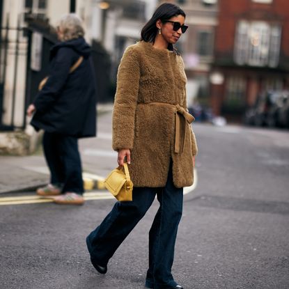 fashion week attendee wearing sunglasses, long faux fur coat, jeans, black boots, and yellow bag