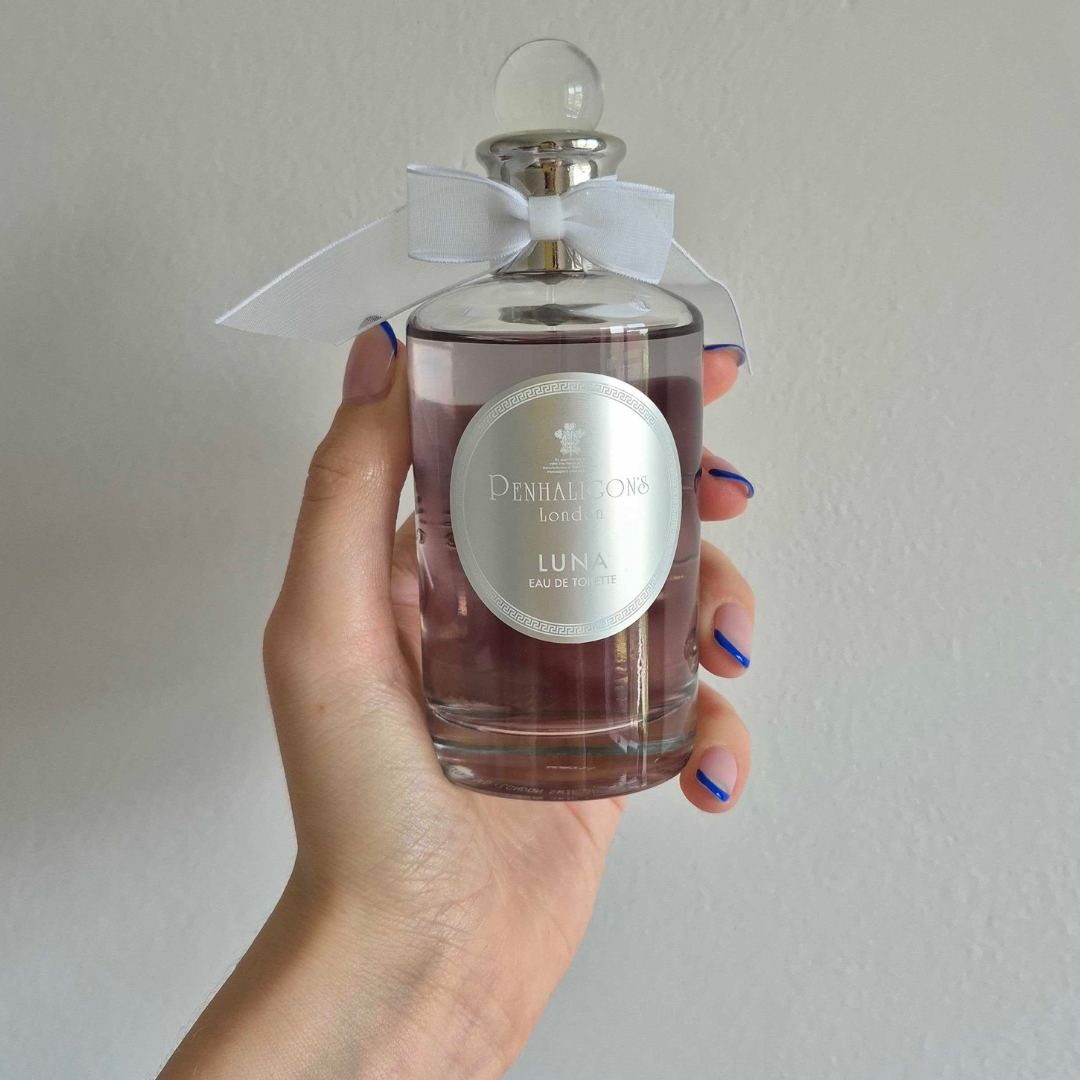 A close-up of Digital Beauty Writer Naomi Jamieson's hand, seen holding a bottle of Penhaligon's Luna perfume, which features a sheer, white bow/ in front of a cream wall background