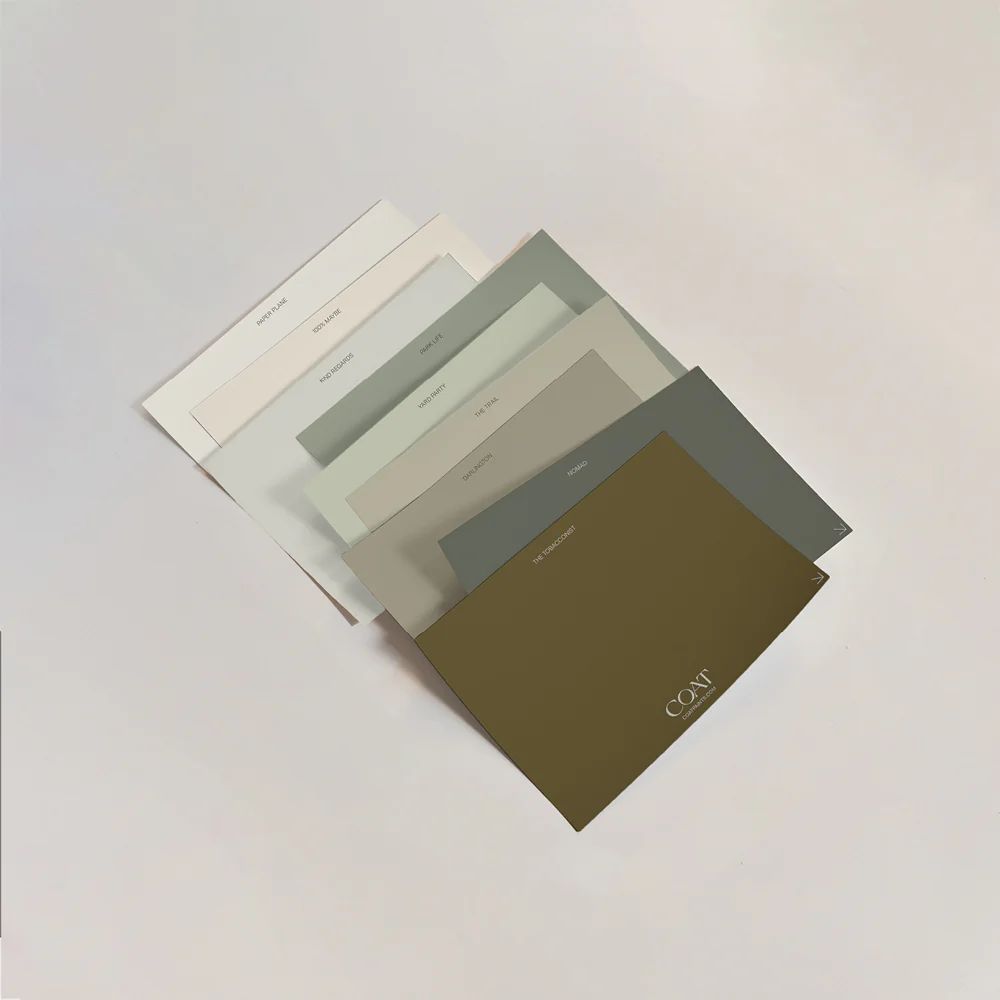 Overlapping paint colour sample cards in soft neutrals and muted greens, arranged on a pale background to show a calm, earthy palette.