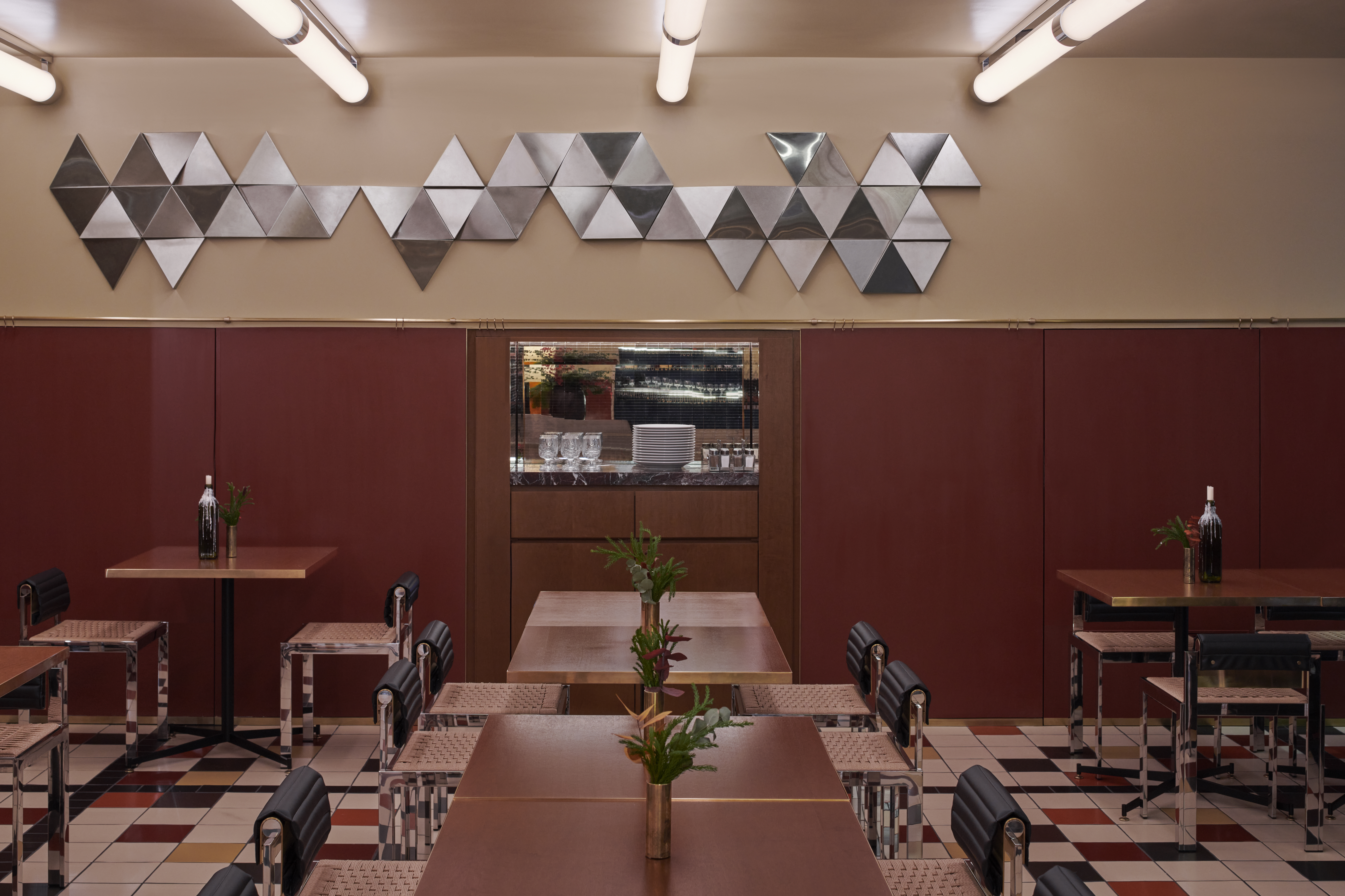 A restaurant&#039;s dining room boasts back-lit coffered ceilings, mid-century furniture in leather, chrome, and wood, framed artworks scattered across its walls, checkered floors, and a downstairs bar clad in brushed steel and golden light.