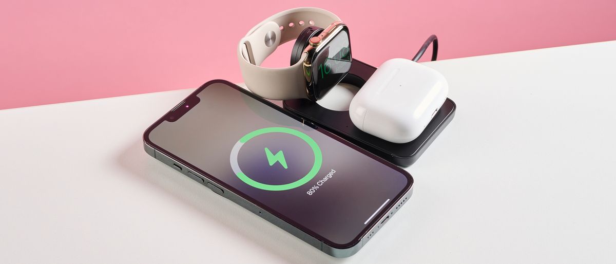 The Mous MagSafe 3 in 1 charger is pictured open on a white tabletop with a pink background. An iPhone, Apple Watch, and Air Pod case are shown charging.