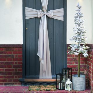 A front door wrapped in a silver ribbon with a snowy potted tree next to it and a Christmas doormat in front of it