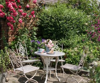 Beautiful English garden-style patio with roses, shrubs and a table and chairs set for afternoon tea