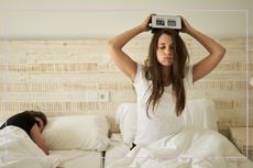 Clock change tips: a tired woman with her eyes shut holding a digital clock above her head sits up in bed beside a sleeping man