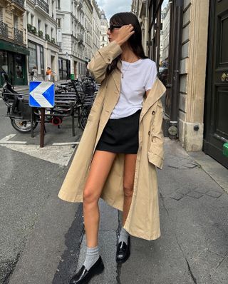 Leia Sfez wearing a trench coat, white T-shirt, black skirt, and loafers with socks in Paris.