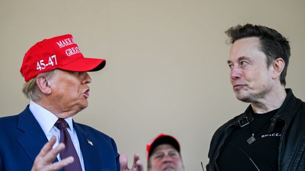 A pale man wearing a blue suit and red hat with orange face paint purses his lips toward another, taller pale man, pursing his lips in the direction of the first man, wearing all black. A blurry man behind them awaits the inevitable.