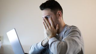 A young man looking at a laptop screen with his hands over his face in shock