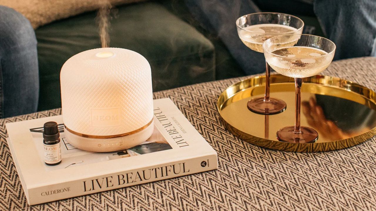 A white Neom Wellbeing Pod Luxe running on a white coffee table book, on a patterned upholstered ottoman. To the right is a gold tray with two brown and clear glass champagne coupes.