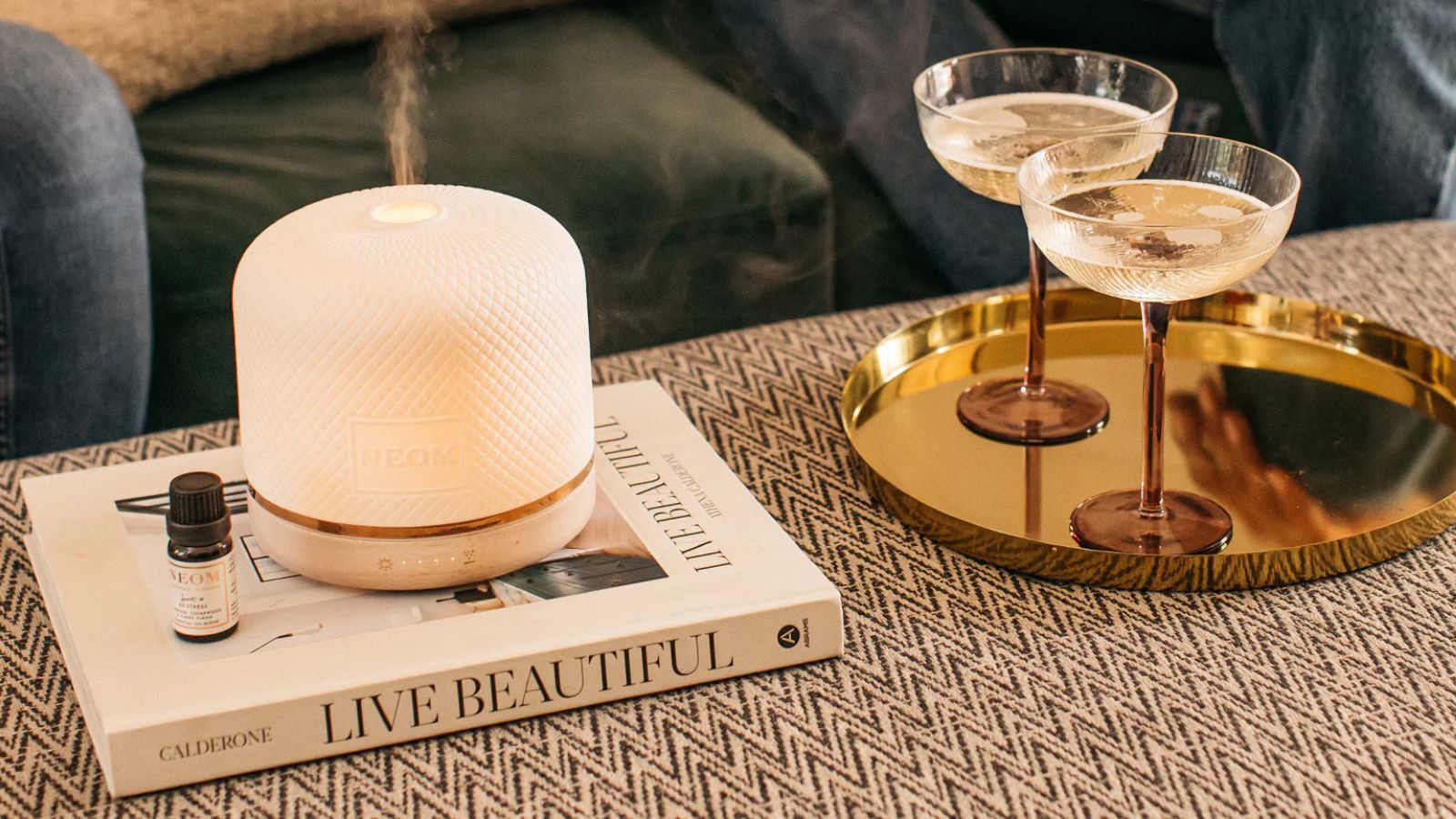 A white Neom Wellbeing Pod Luxe running on a white coffee table book, on a patterned upholstered ottoman. To the right is a gold tray with two brown and clear glass champagne coupes.
