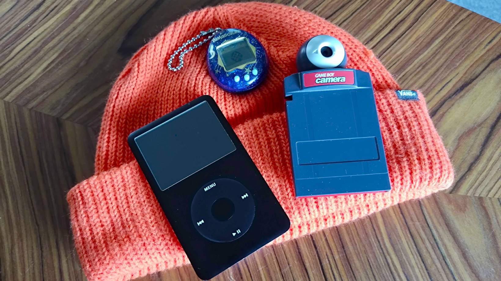 iPod Classic, Game Boy Camera, and Blue Tamagotchi sitting on orange hat.
