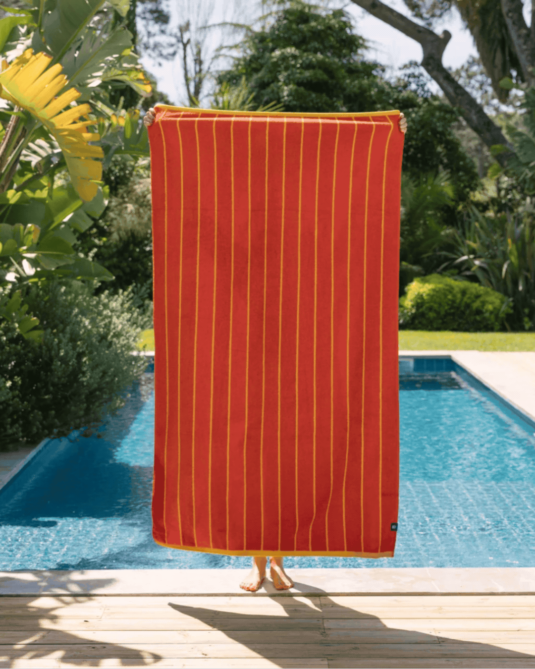 A striped red, ochre, and pale blue beach towel, held vertically by a person hiding behind it in front of a sun-lit pool.