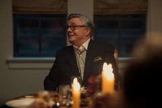 Nathan Lane as Dominick Dunne, sitting at a dinner table behind lit candles, in episode 208 of Monsters: The Lyle And Erik Menendez Story.