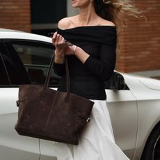 Woman carrying brown suede bag and wearing off-the-shoulder dress with black knit top and flowing white skirt.