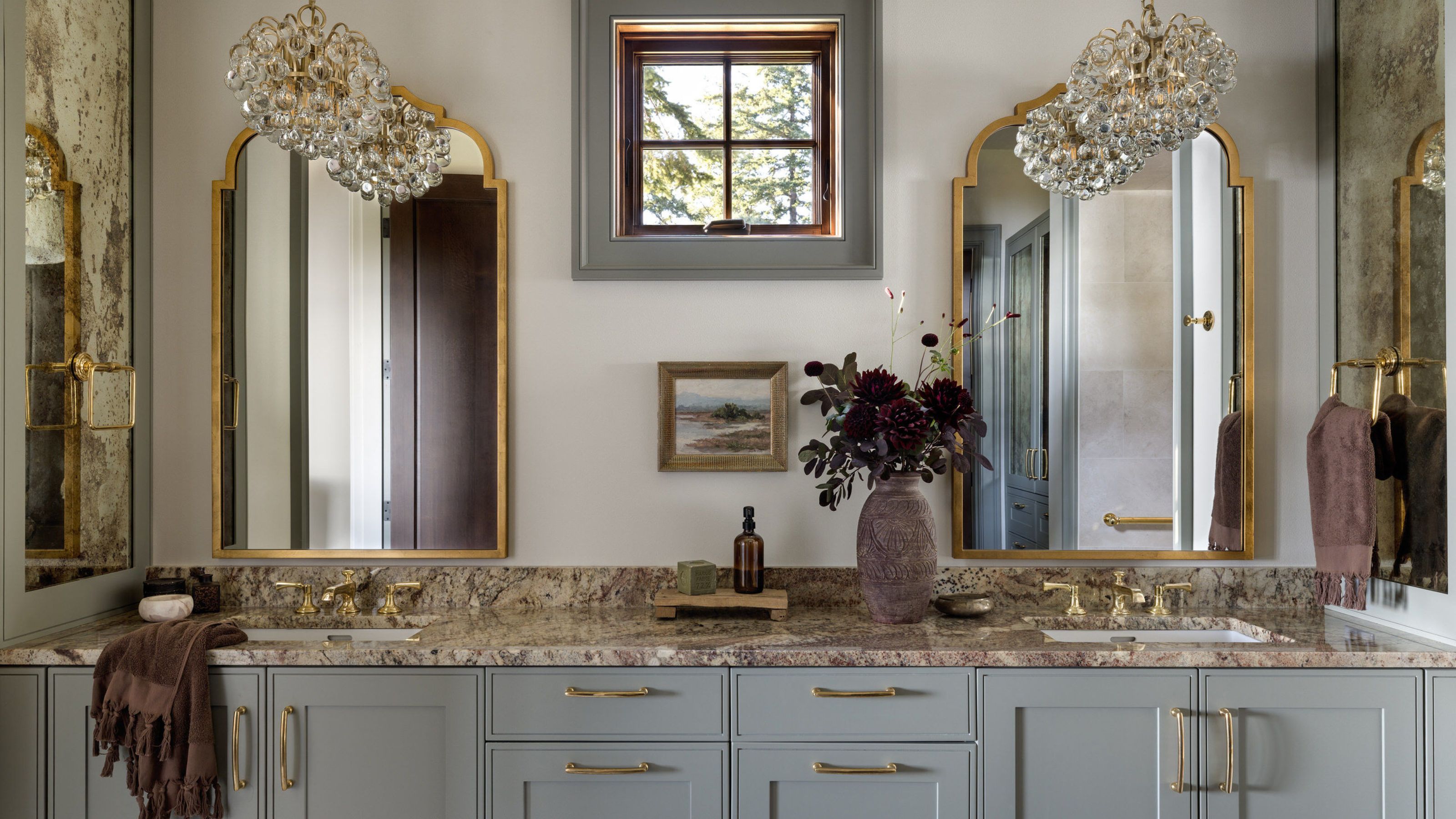 A sophisticated bathroom with warm white walls, pale blue cabinets, marble countertops, two brass mirrors on the wall, brass fixtures, and two glass pendant lights.